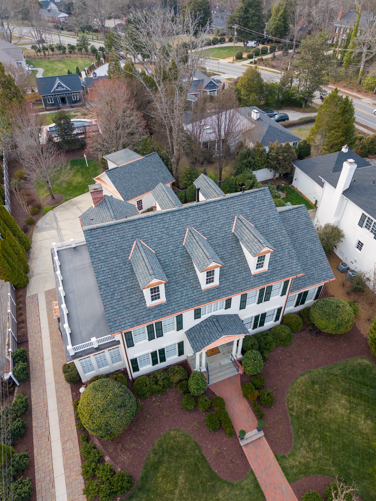 Aerial view of a large two-story house with gray roof, white exterior, and green shutters, surrounded by manicured lawns, shrubs, and brick walkways in a suburban neighborhood.