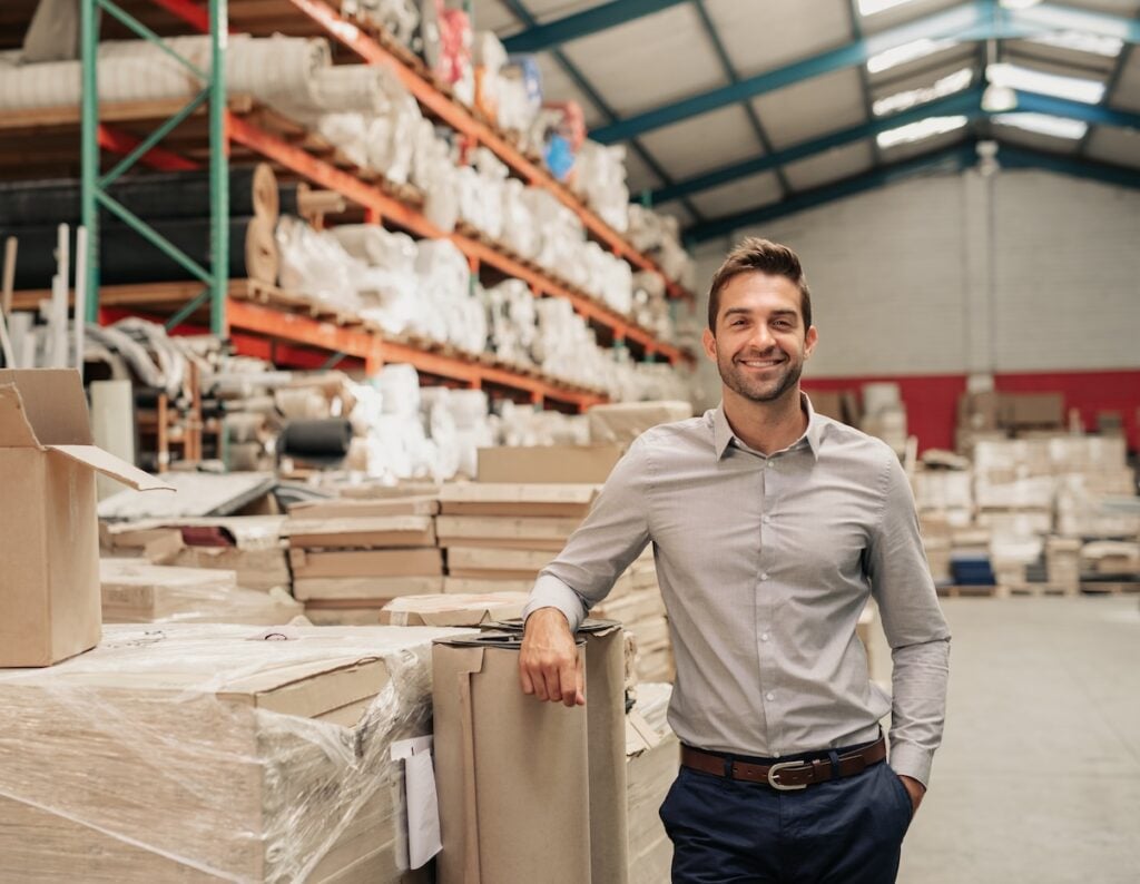 Portrait of a smiling warehouse manager leaning against some stock with piles of carpets stacked on shelves in the background