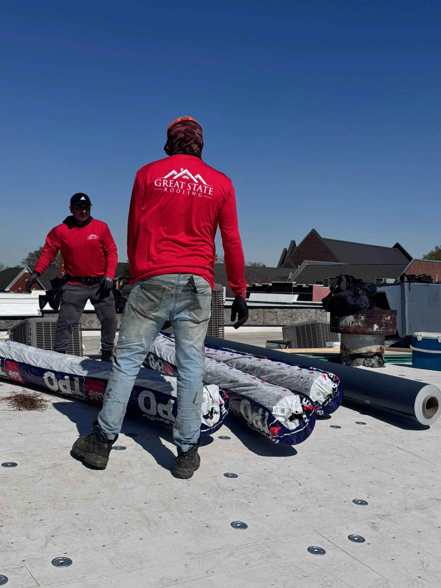 Two workers in red shirts and jeans stand on a residential roof under a clear blue sky, carrying a large roll of roofing material. Roofing supplies and tools are scattered around them, with nearby houses visible in the background.