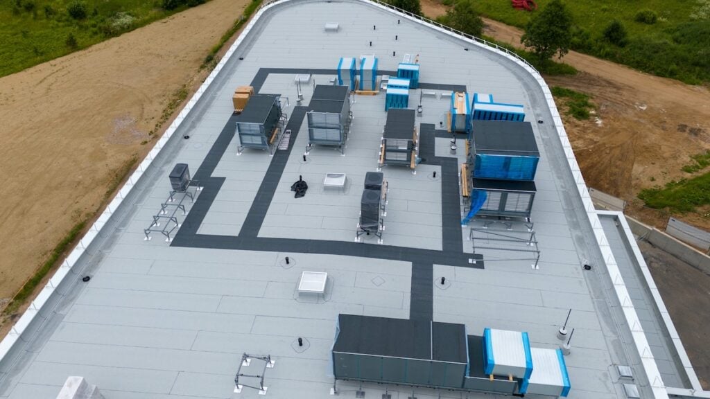 Aerial view of a commercial building rooftop featuring various HVAC units, ventilation systems. The flat roof is mostly gray with black pathways.
