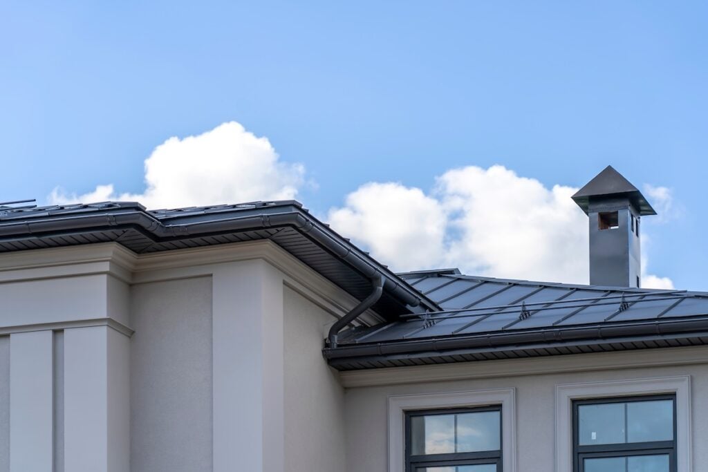 view of house with gray fold roof and plums and filing of roof overhangs with soffits and brick stone pipe covered with metal sheets and black smoke box