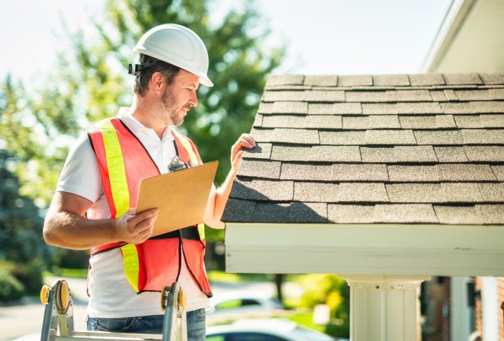 roof inspection cost A man with hard hat standing on steps inspecting house roof