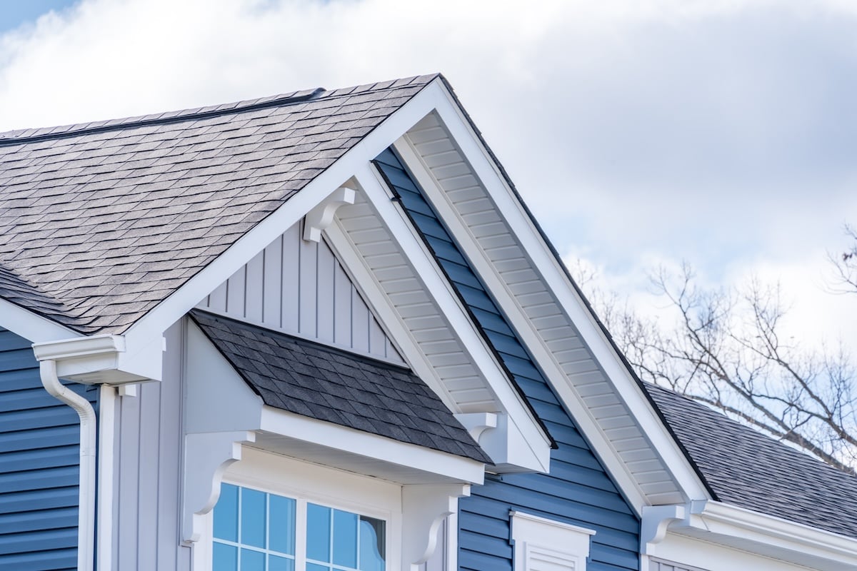 Elegant American house facade, with Shed Roof Over Awning Window, double gable white brackets, gray vertical and blue horizontal vinyl lap siding, white fascia with cloudy blue sky