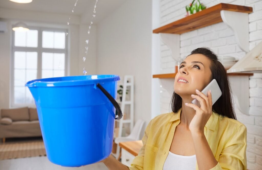 ceiling leaking in apartment Sad unhappy frustrated young housewife lady looking up at water falling down from damaged ceiling in newly renovated home interior, holding pail and speaking to repair service. House roof leak concept
