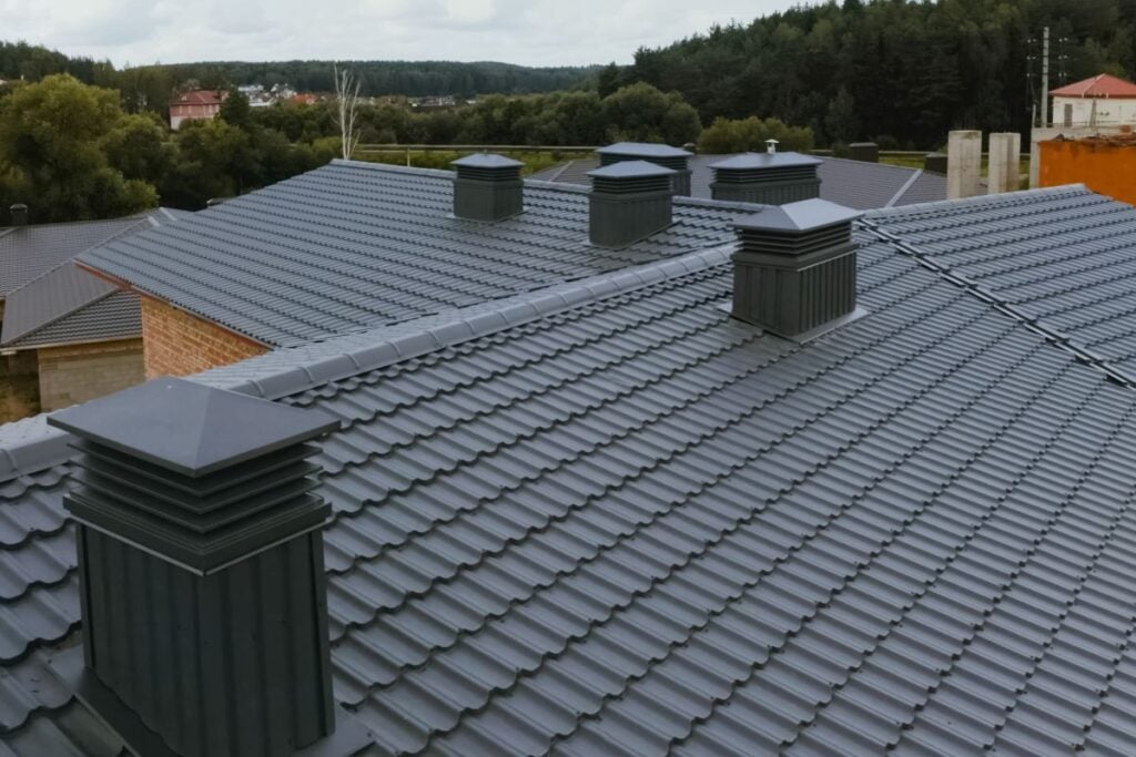 burnished slate metal roofing Multiple gray burnished slate metal rooftops with large square ventilation chimneys, set among trees and other buildings in the background, under a cloudy sky.