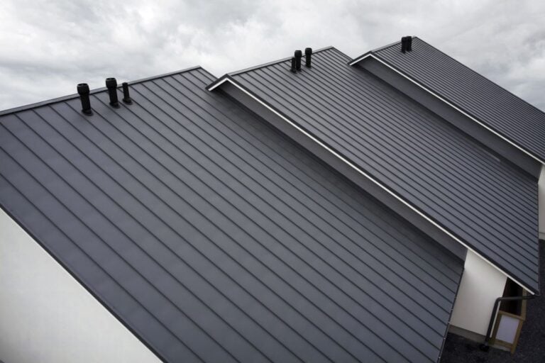 burnished slate metal roofing Four modern slanted rooftops feature sleek Burnished Slate Metal Roofing panels and several vertical chimneys, set against a cloudy sky. The roofs are arranged in a row, creating a striking geometric pattern.