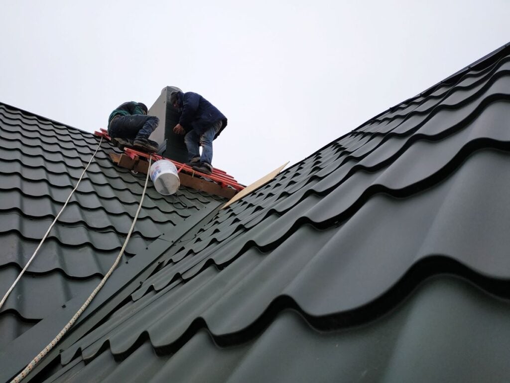 burnished slate metal roofing Two workers in safety gear repair or install trending metal roofing in Burnished Slate on a steep building slope, with ropes, tools, and a bucket nearby on a cloudy day.