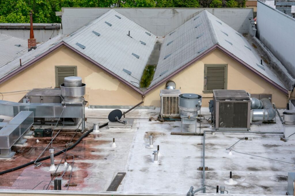 commercial roof financing Rooftop with various HVAC units, vents, ducts, and a satellite dish sits atop a sloped roof with louvered vents; trees are visible in the background—ideal for exploring Commercial Roof Financing to fund repairs or upgrades.