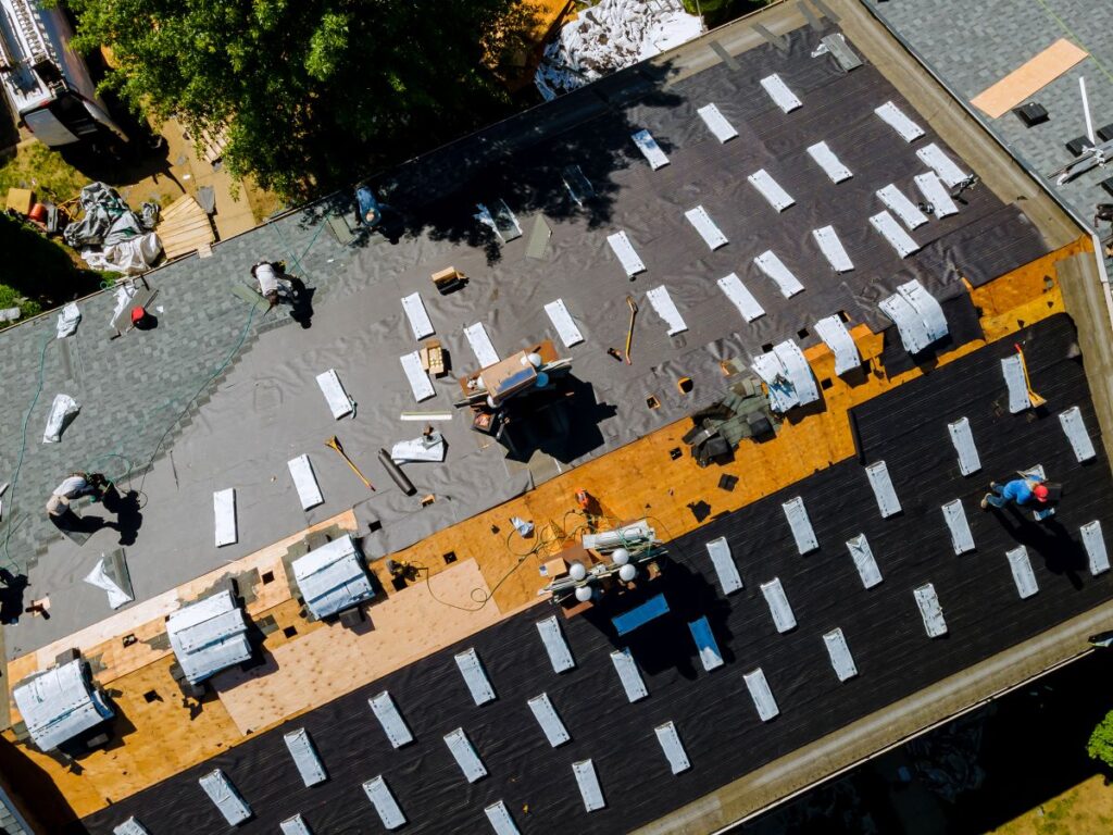 commercial roof financing Aerial view of workers installing new roofing on a large building, with tools and materials neatly organized. Some areas show black underlayment, others new shingles—ideal for those considering commercial roof financing or roof repairs.