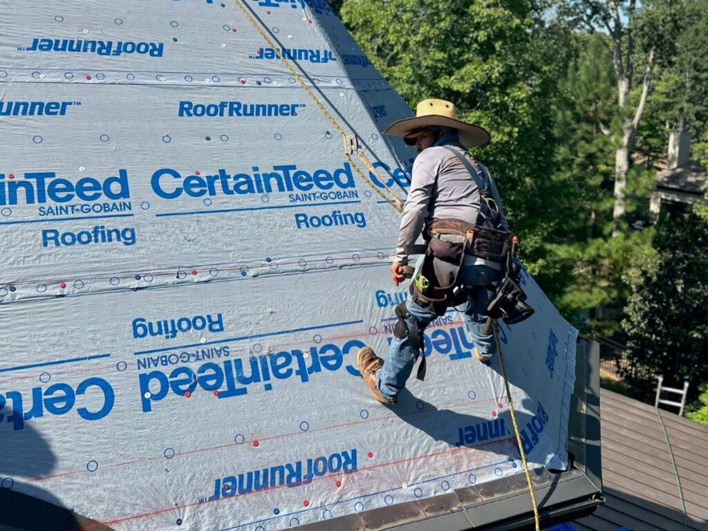 commercial roof snow removal worker wearing safety gear and a wide-brim hat is installing CertainTeed roofing underlayment on a steep roof, surrounded by trees on a sunny day—promoting winter roof safety best practices.