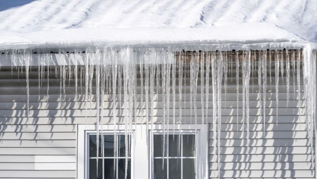 commercial roof snow removal Long, clear icicles hang from the snowy edge of a roof above a white siding house with double glass doors, casting vertical shadows on the wall in bright sunlight—a reminder of the importance of winter roof safety.