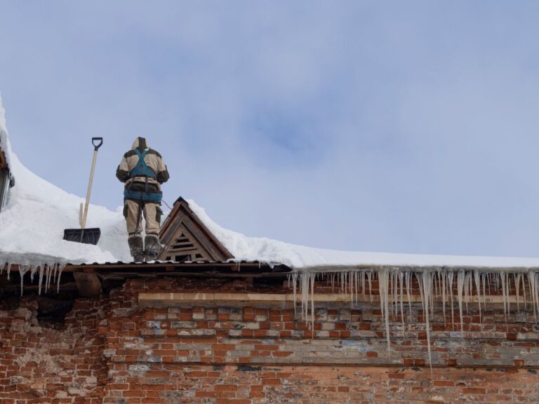 commercial roof snow removal person in winter gear stands on a snow-covered roof next to a shovel, demonstrating winter roof safety as icicles hang from the edge of the old brick building under a cloudy sky.