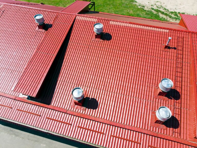 commercial roof financing Aerial view of a large red metal roof with several circular vents and a small white antenna, ideal for Roof Repairs. Green grass and sandy ground are visible around the building.