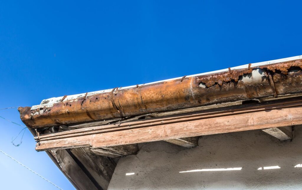 commercial gutter repair close-up view of a rusted, damaged commercial gutter system attached to the edge of a roof against a clear blue sky. The gutter shows significant corrosion and deterioration, which are clear gutter repair signs.