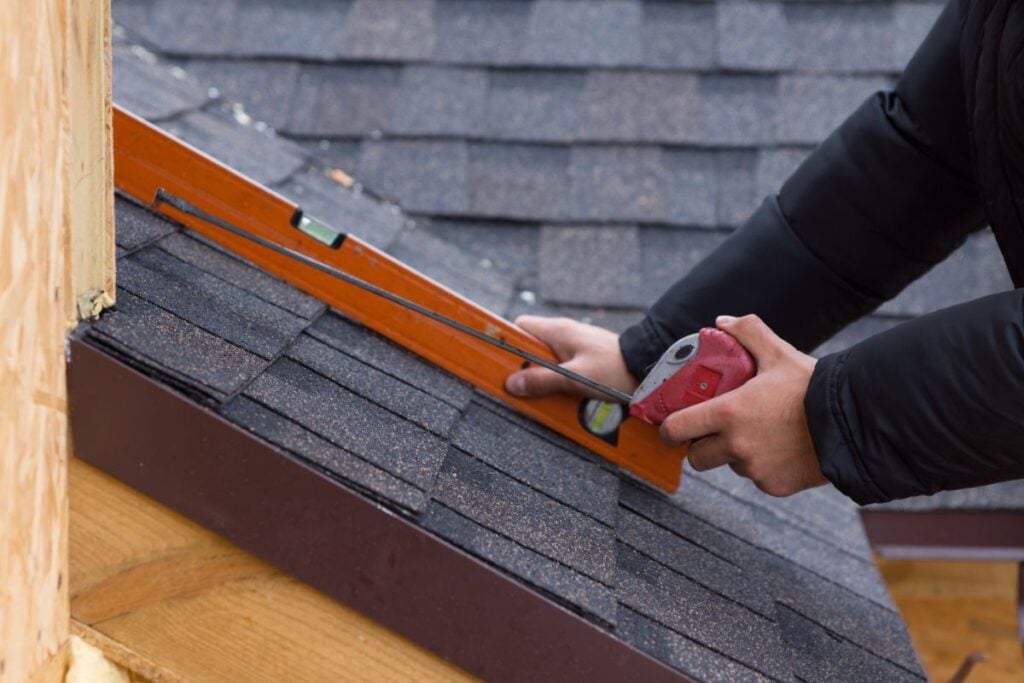 how to measure a roof for metal roofing Person using a spirit level and a measuring tape to check the alignment of roof shingles—an essential part of the roofing steps when you measure a roof on a house under construction.