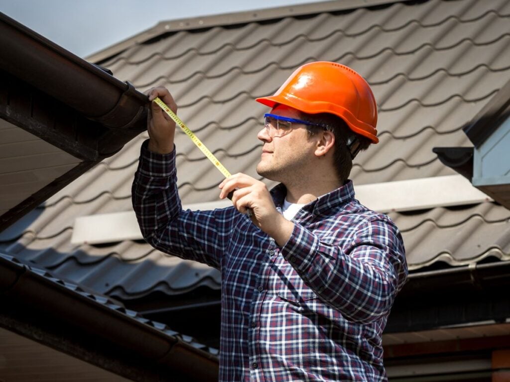 how to measure a roof for metal roofing person wearing a hard hat and safety glasses stands on a ladder, using a tape measure to perform a roof measurement on the gutter of a house with metal roofing. Tools are clipped to their belt.