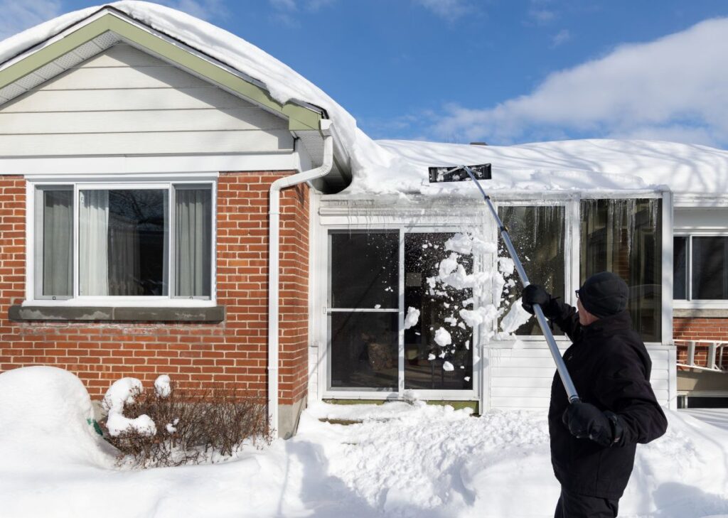 commercial roof snow removal person in winter clothing uses a long-handled tool for roof snow removal, clearing heavy snow from the house roof. The yard and surrounding area are blanketed in white under a blue sky, highlighting the importance of winter roof safety.