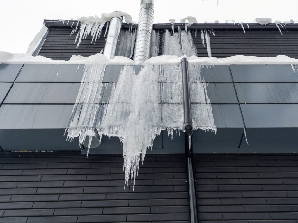 industrial roof insulation Large icicles hang from the edge of a modern dark building’s roof, highlighting the benefits of industrial roof insulation in cold climates, with snow piled on top and metal pipes extending upward.