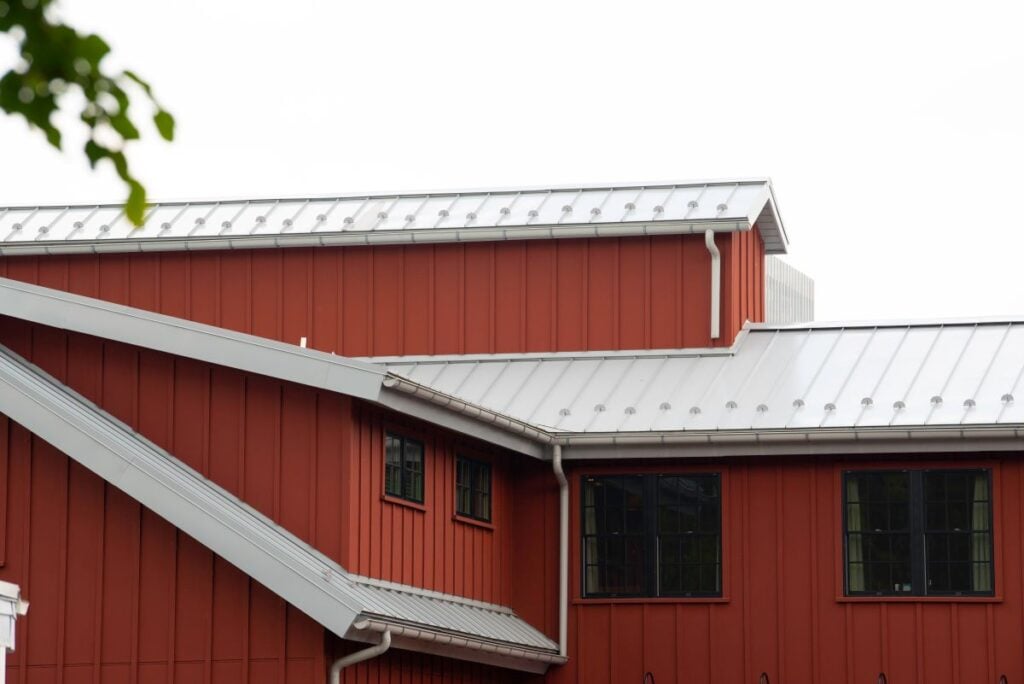 low slope metal roofing Red barn-style building with vertical siding and black-trimmed windows, featuring multiple low slope metal roofs under a bright, overcast sky. A leafy branch is partially visible in the upper left corner.