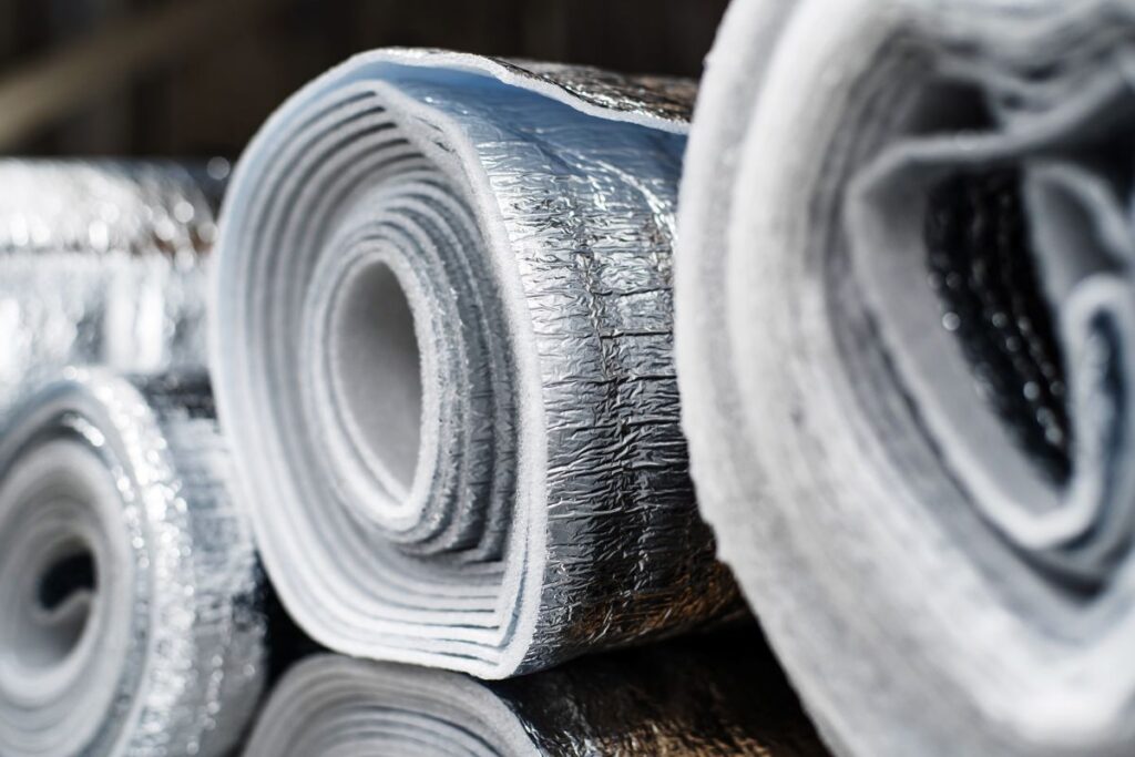 industrial roof insulation Close-up of several rolls of reflective insulation material with metallic surfaces, positioned on a shiny surface. The texture and layers of the insulation are clearly visible.