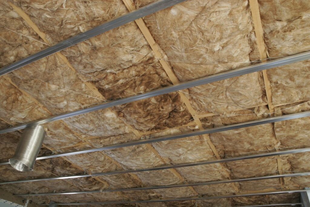 industrial roof insulation ceiling with exposed fiberglass insulation between wooden beams and several metal support beams, along with a silver ventilation duct visible on the left side.