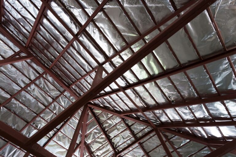 industrial roof insulation View of a roof structure with exposed metal beams and shiny silver insulation panels covering the underside of the roof. The angular beams form geometric patterns against the reflective surface.