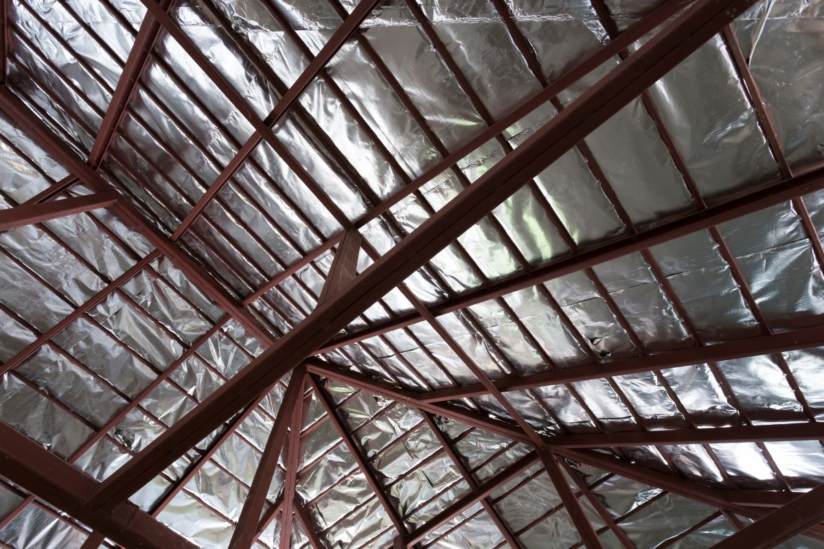 industrial roof insulation View of a roof structure with exposed metal beams and shiny silver insulation panels covering the underside of the roof. The angular beams form geometric patterns against the reflective surface.