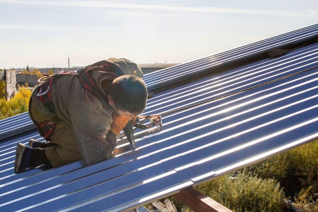 low slope metal roofing worker wearing safety gear kneels on a sloped metal roof, installing or securing panels with a power tool, under bright sunlight with greenery below—demonstrating expertise in Low Slope Metal Roofing.