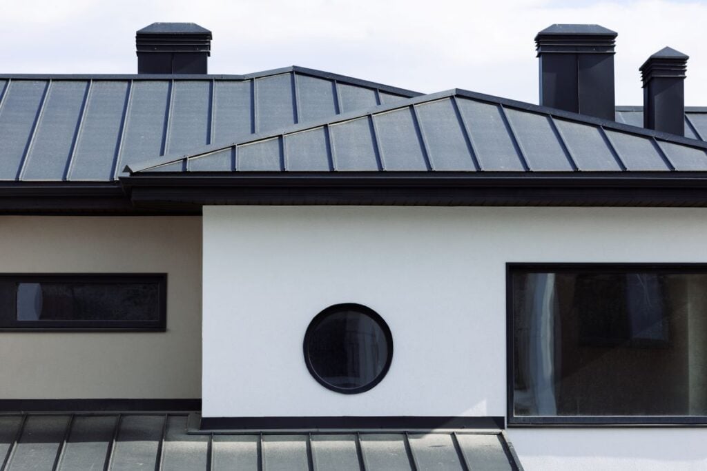 pvdf coating Modern house with a PVDF coating on its metal roof, white and beige exterior walls, a rectangular window on the right, and a small round window in the center. Two black chimneys rise against a cloudy sky.
