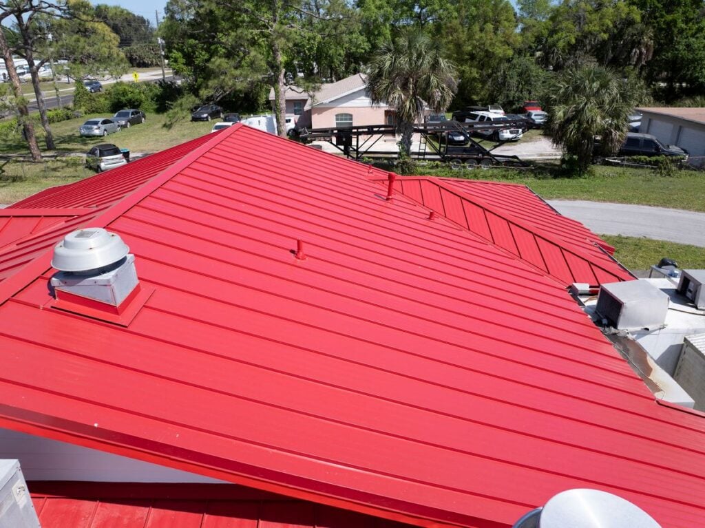 low slope metal roofing Bright red metal roofing systems cover the building, viewed from above, with vents and chimneys visible; the area features parked cars, palm trees, greenery, and nearby roads under a sunny sky. Ideal for low slope metal roofing or flat roofs.
