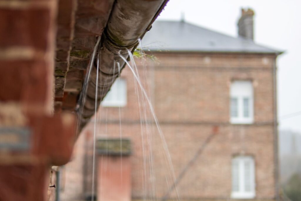 commercial gutter repair close-up of a leaking gutter system on a brick building, with streams of water pouring out—a clear example of gutter repair signs. In the background, another brick building with white-framed windows is slightly out of focus.