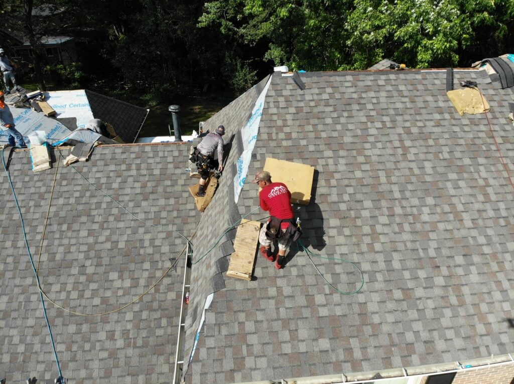 asphalt roof repair Two workers with safety gear perform asphalt roof repair, installing or fixing shingles on a house roof using wooden boards and tools. Green trees and other rooftops are visible in the background.