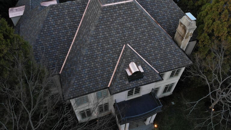 asphalt roof rejuvenation Aerial view of a large house with a dark shingle roof, copper flashing, and white exterior walls. Surrounded by trees, this home could benefit from asphalt roof rejuvenation to help extend roof life and maintain its impressive appearance.