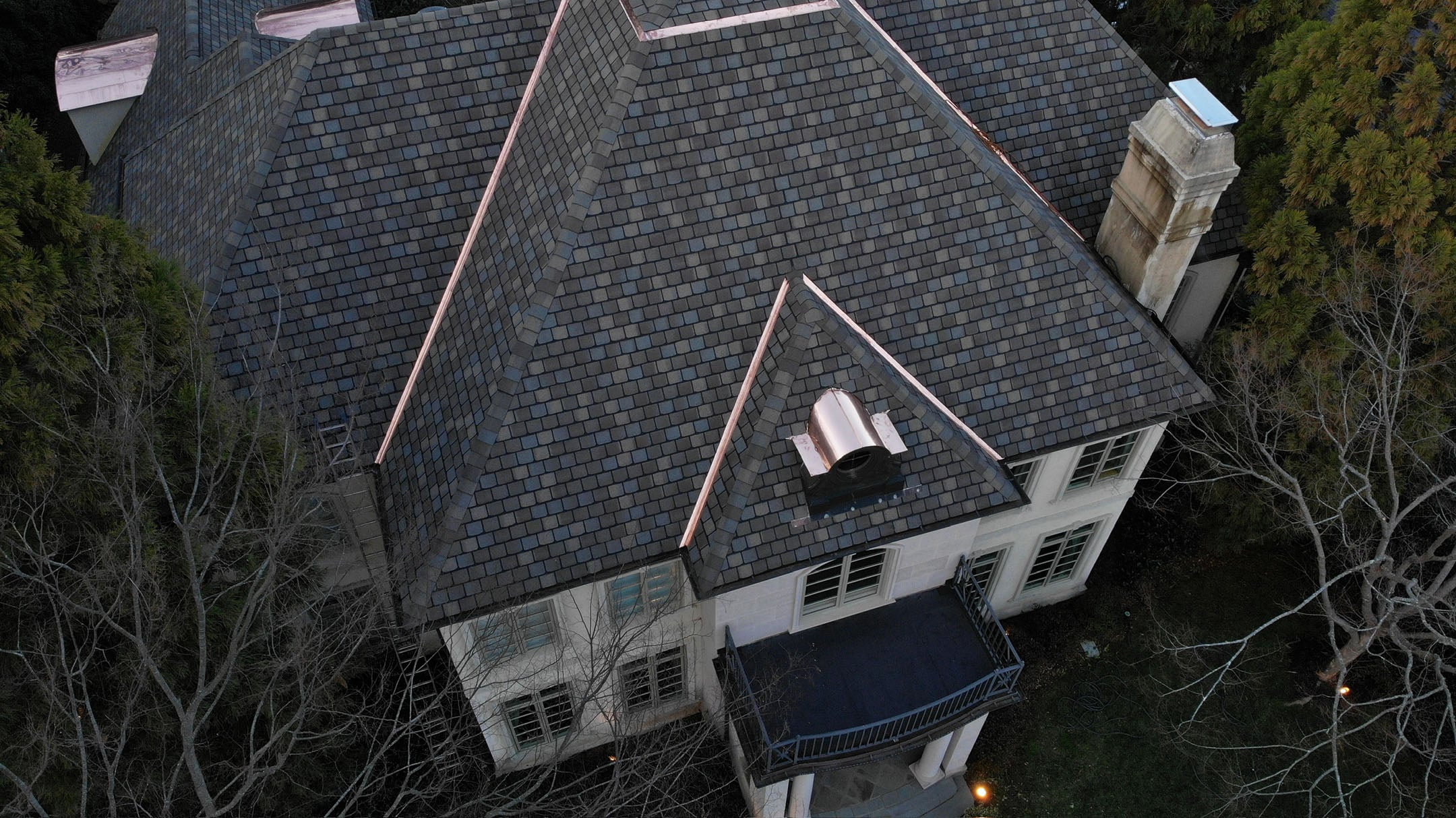 asphalt roof rejuvenation Aerial view of a large house with a dark shingle roof, copper flashing, and white exterior walls. Surrounded by trees, this home could benefit from asphalt roof rejuvenation to help extend roof life and maintain its impressive appearance.