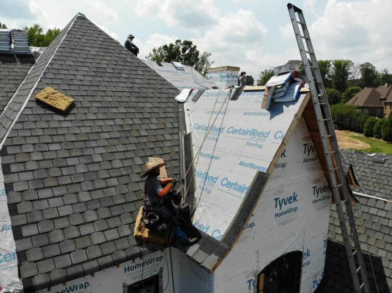 asphalt roof repair A worker wearing a hat and tool belt installs asphalt roofing shingles on the steep roof of a house under construction. The roof is partially covered with protective wrap, and a ladder leans against the house. Other workers are visible in the background.