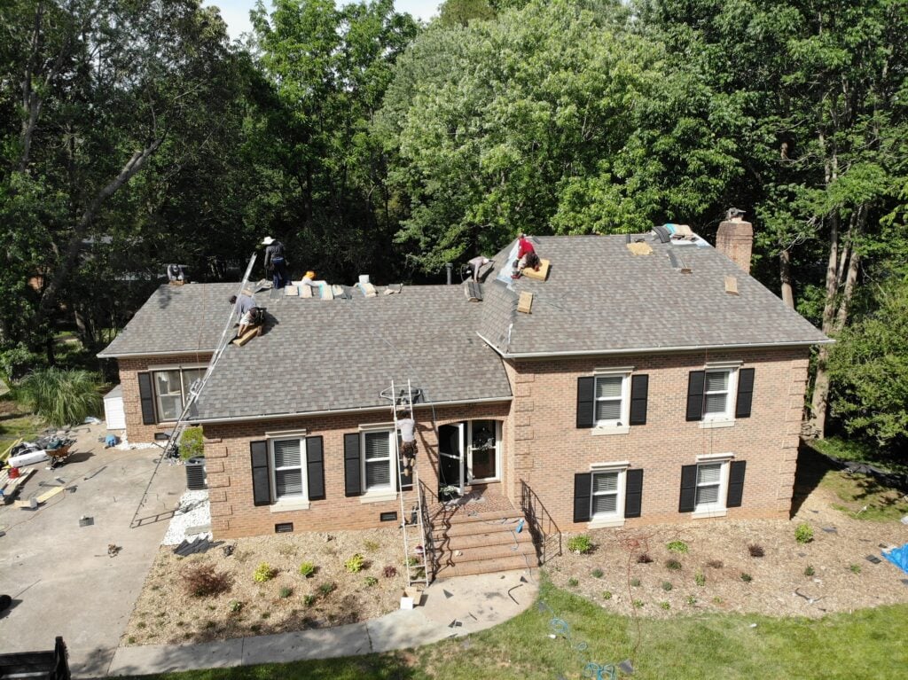 asphalt roof rejuvenation Aerial view of workers repairing the roof of a two-story brick house surrounded by trees, with ladders and construction materials visible—demonstrating asphalt roof rejuvenation to extend life and protect this home.