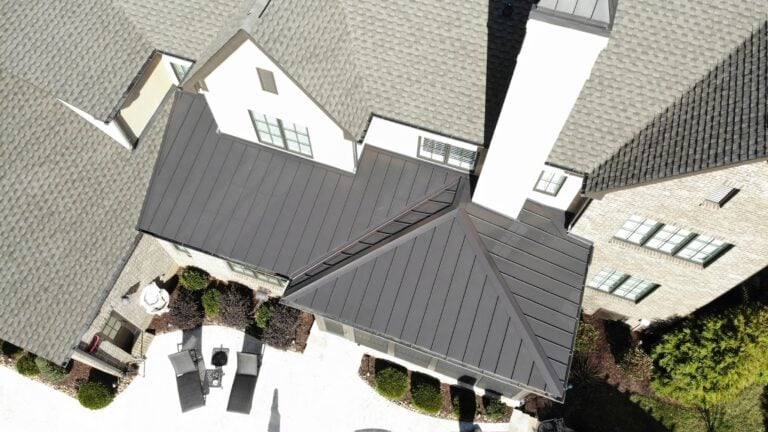 alternative to asphalt roofing Aerial view of a house with a gray shingled roof, metal-roofed section, and white chimney. Patio furniture and landscaping are visible in the backyard, highlighting attractive alternative to asphalt roofing options for 2026.