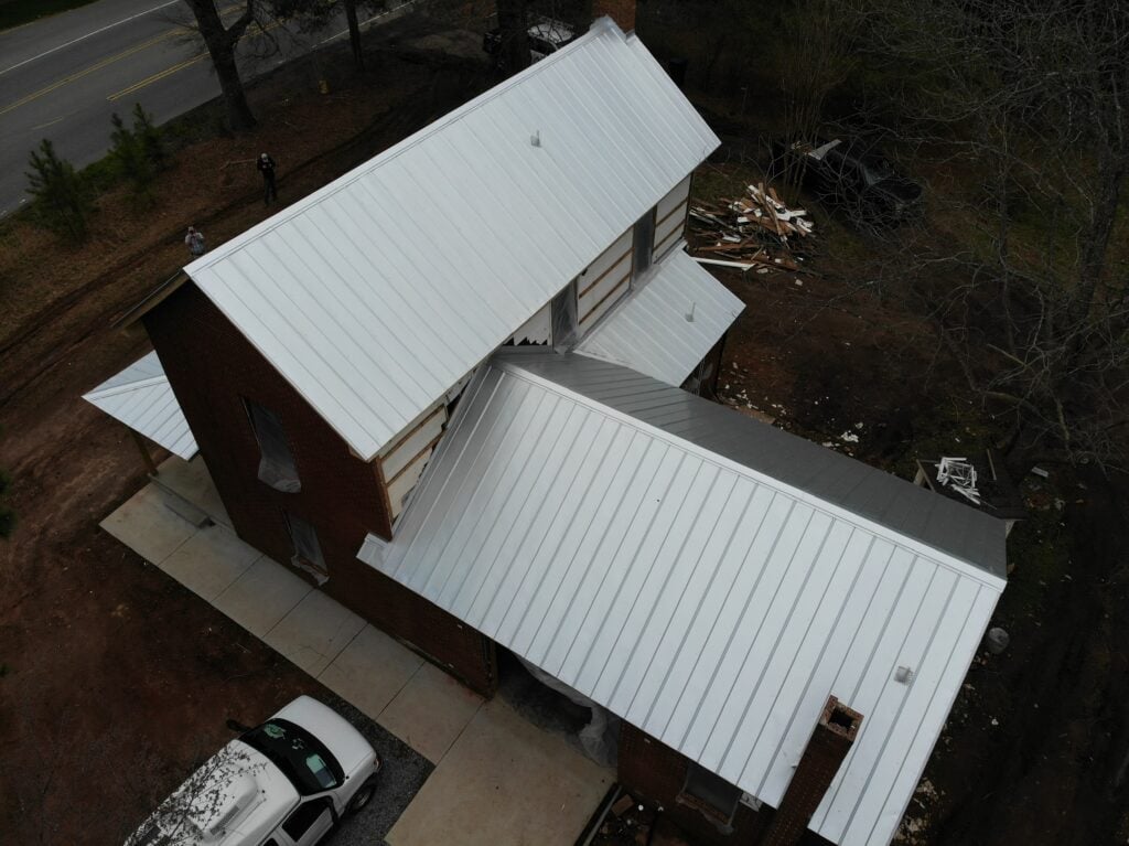metal roof vs asphalt shingles Aerial view of a brick house with a white metal roof, highlighting differences from asphalt shingles. Surrounded by bare trees and construction materials, a white vehicle is parked on the driveway near the house, with a road running nearby.