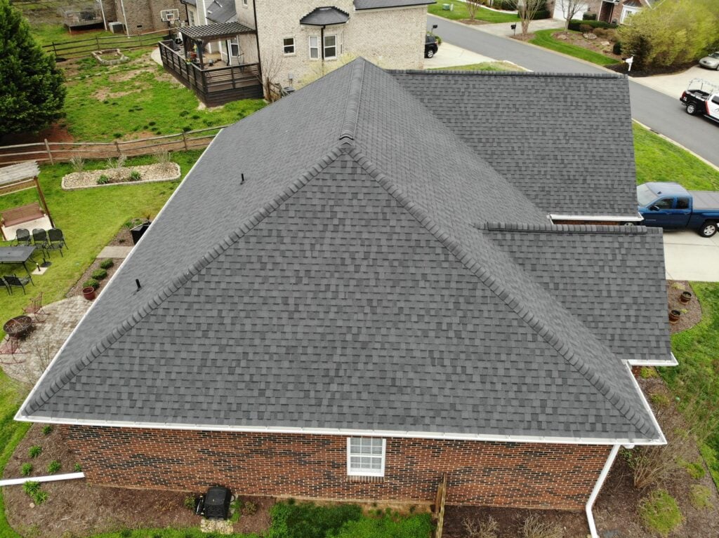metal roof vs asphalt shingles Aerial view of a house with a large, dark gray asphalt shingles roof and red brick exterior, surrounded by a green lawn, patio area, and nearby houses—highlighting roof differences in the neighborhood.
