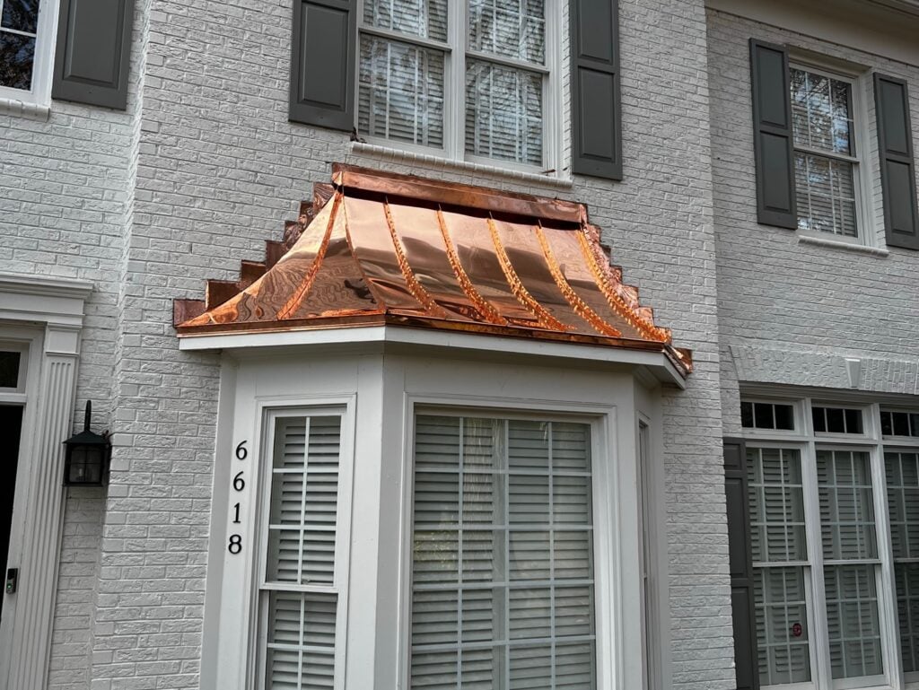 alternative to asphalt roofing A two-story house with light gray brick exterior, gray shutters, and a bay window covered by a shiny copper roof—an elegant alternative to asphalt roofing. The house number 6618 is displayed vertically beside the window.
