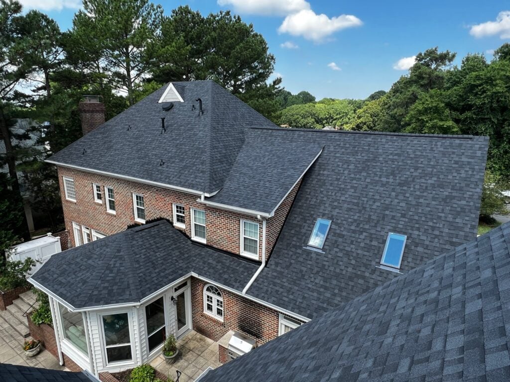 alternative to asphalt roofing A large brick house with multiple roof sections covered in dark gray shingles, several windows, and skylights, surrounded by tall trees and greenery under a blue sky—an inspiring example of alternative to asphalt roofing.