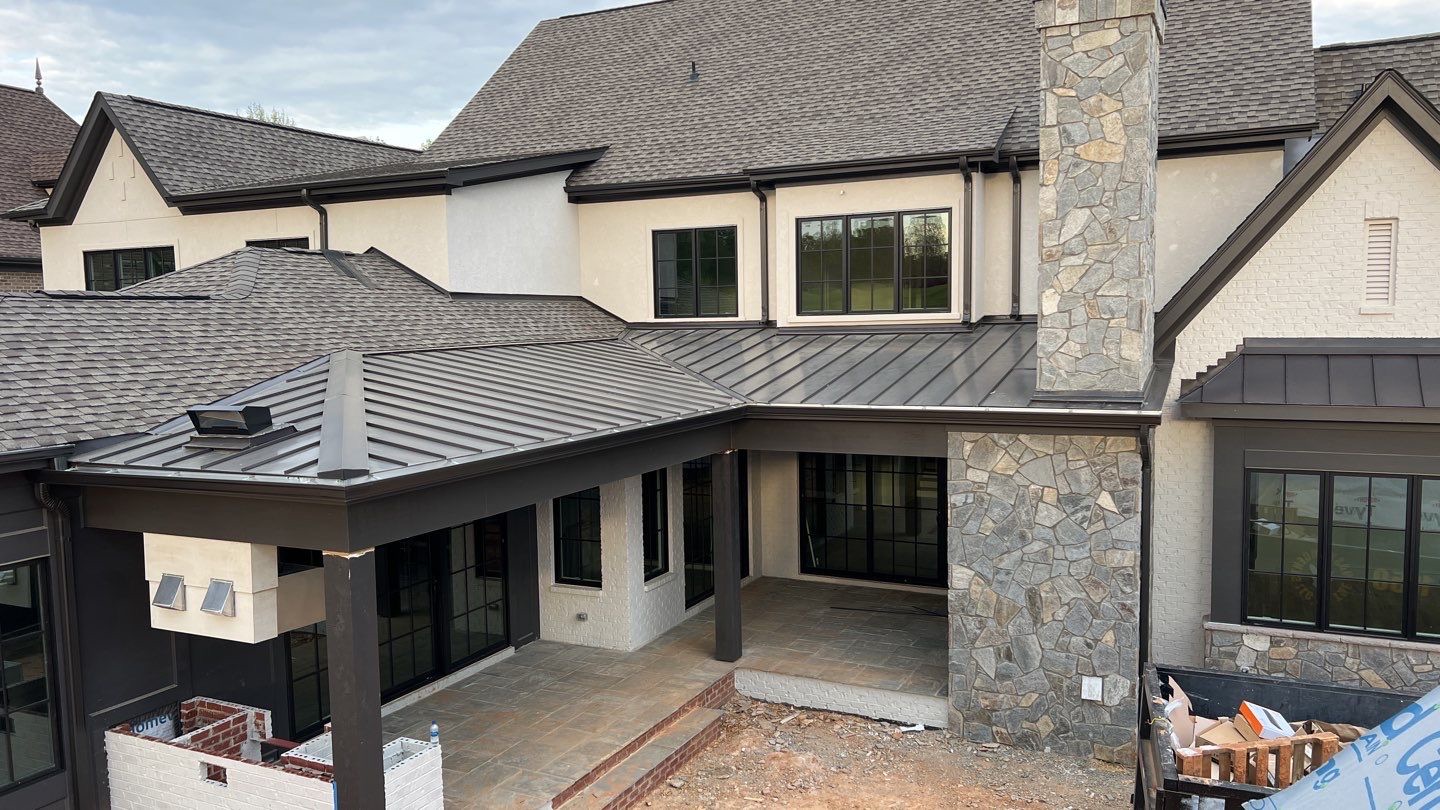 metal roof vs asphalt shingles A modern two-story house under construction, featuring large black-framed windows, a stone chimney, a covered porch with a metal roof highlighting roof differences from homes with asphalt shingles, light-colored exterior walls, and an unfinished yard area.