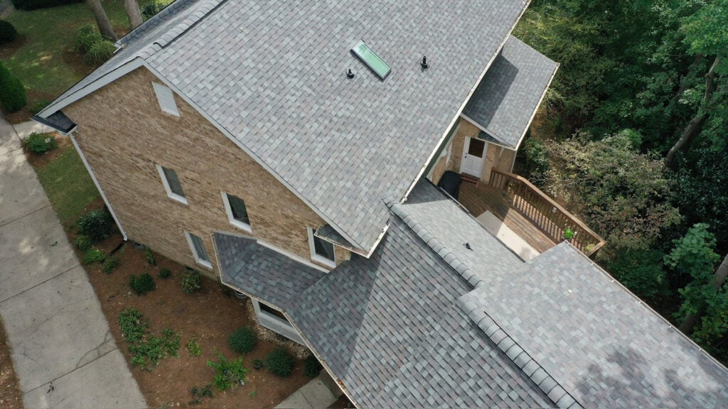 alternative to asphalt roofing Aerial view of a two-story brick house with a gray asphalt shingle roof, a skylight, wooden deck, and lush trees—showcasing the classic charm of asphalt shingles in the ongoing metal roof vs asphalt shingles debate.