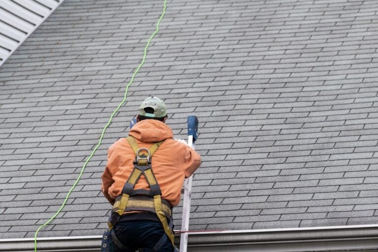 asphalt roof maintenance A person wearing a harness and orange hoodie climbs a ladder to reach the roof of a house with gray shingles, using a green safety rope—a smart move for anyone following asphalt roof maintenance tips.