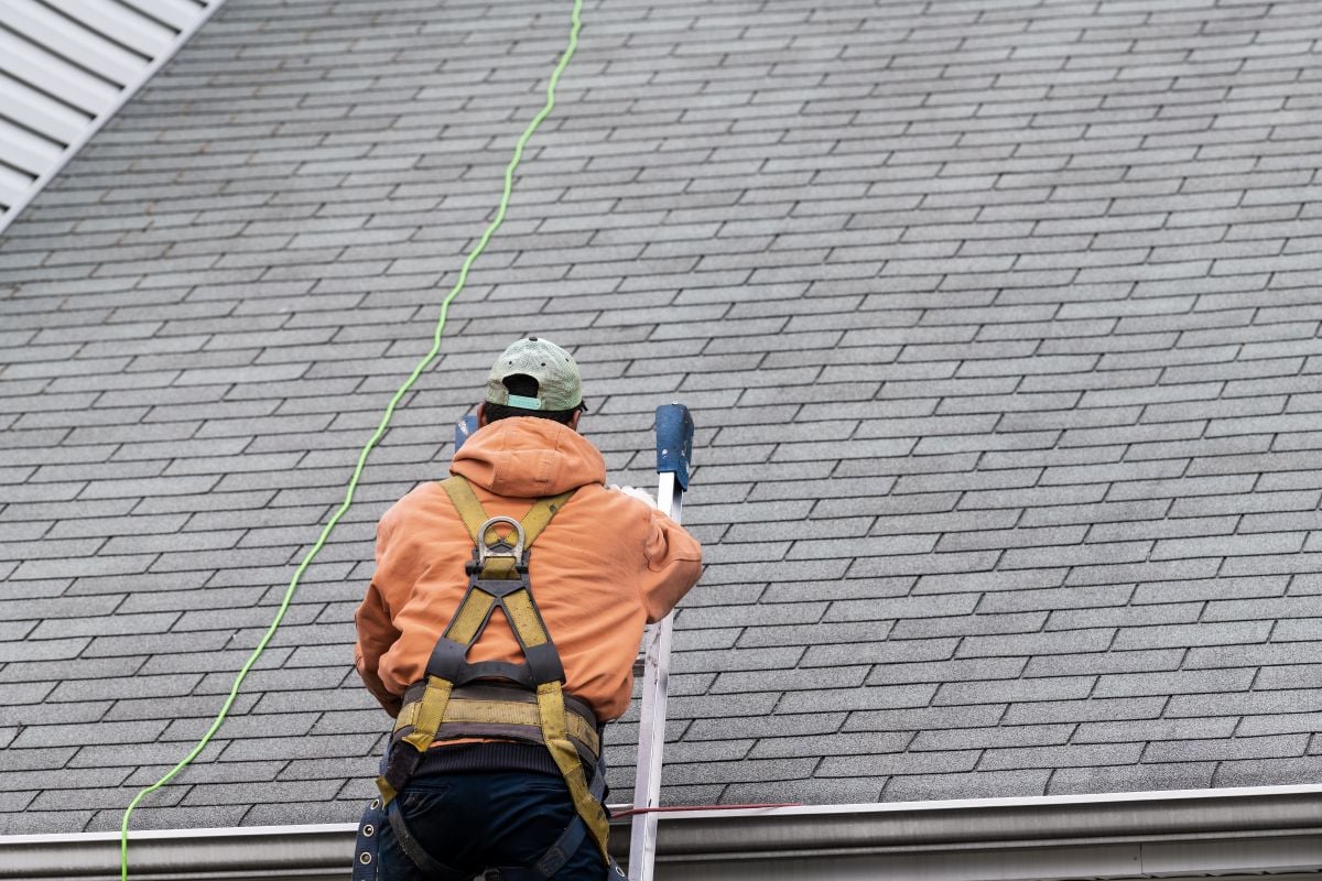 asphalt roof maintenance A person wearing a harness and orange hoodie climbs a ladder to reach the roof of a house with gray shingles, using a green safety rope—a smart move for anyone following asphalt roof maintenance tips.