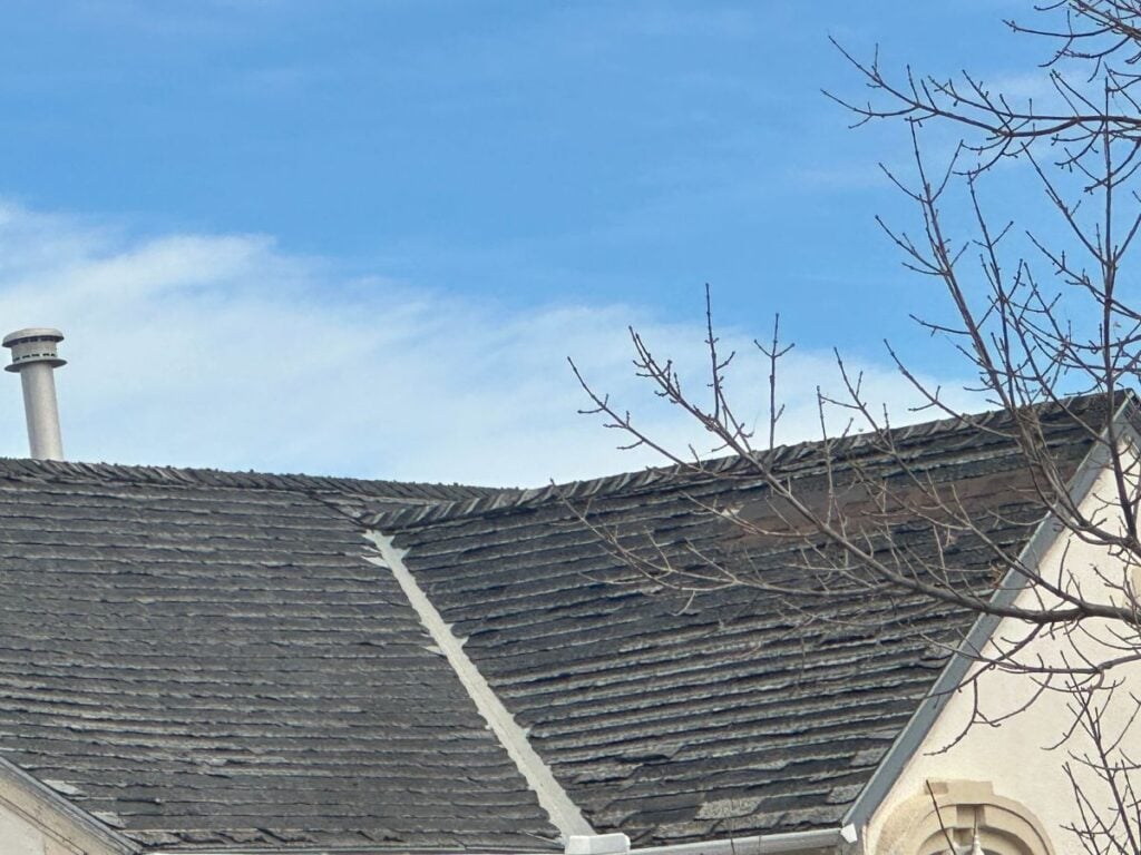 asphalt roof rejuvenation A close-up view of a weathered rooftop with dark shingles hints at the need for asphalt roof rejuvenation to extend life, featuring a small chimney, part of a beige building, and bare tree branches against a blue sky with some clouds.