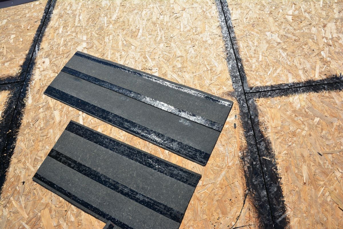 asphalt roofing cement Two asphalt roofing shingles with black adhesive strips lie on a wooden roof deck, which has black tar lines outlining sections, showing the uses and benefits of asphalt roofing cement in preparing for shingle installation.
