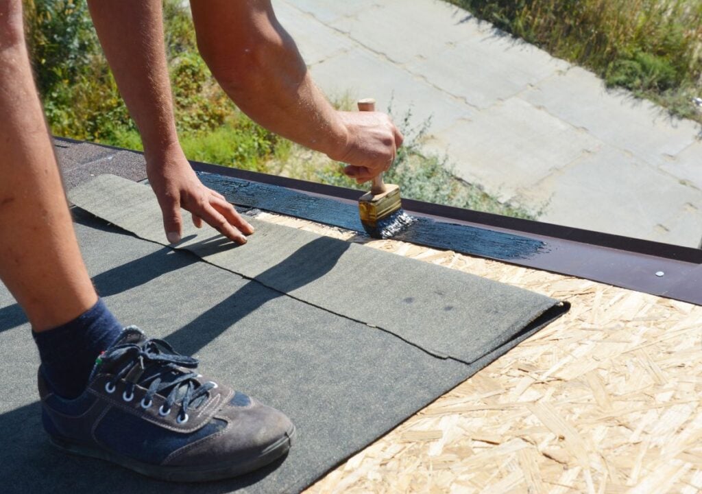 asphalt roofing cement A person applies asphalt roofing cement with a brush to the edge of a roof while standing on plywood and wearing sneakers, demonstrating one of the common uses and benefits of this material for sealing and repair.