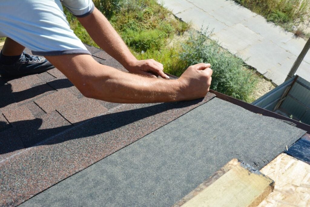 asphalt roofing cement A person installs asphalt shingles on a roof, skillfully using their hands and applying asphalt roofing cement to secure the shingles. The scene is outdoors, with surrounding greenery and a partial sidewalk visible below.