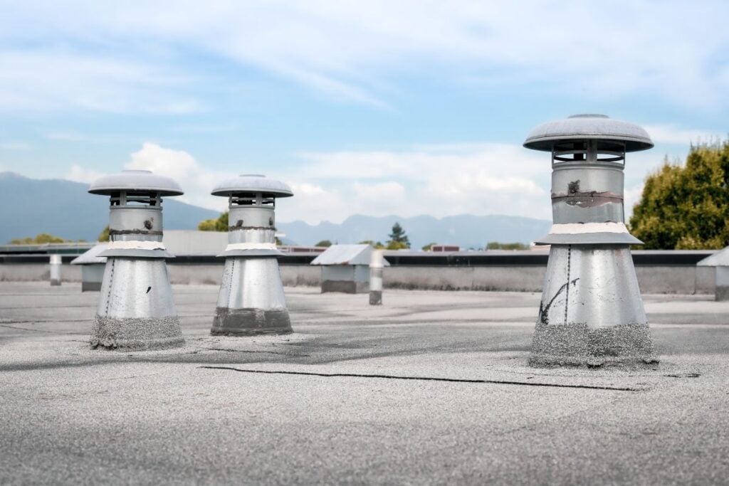 cap sheet Three metal rooftop vents with cylindrical bases and domed tops are positioned on a cap sheet surface, typical for flat roofs, against a backdrop of blue sky, clouds, trees, and distant mountains.